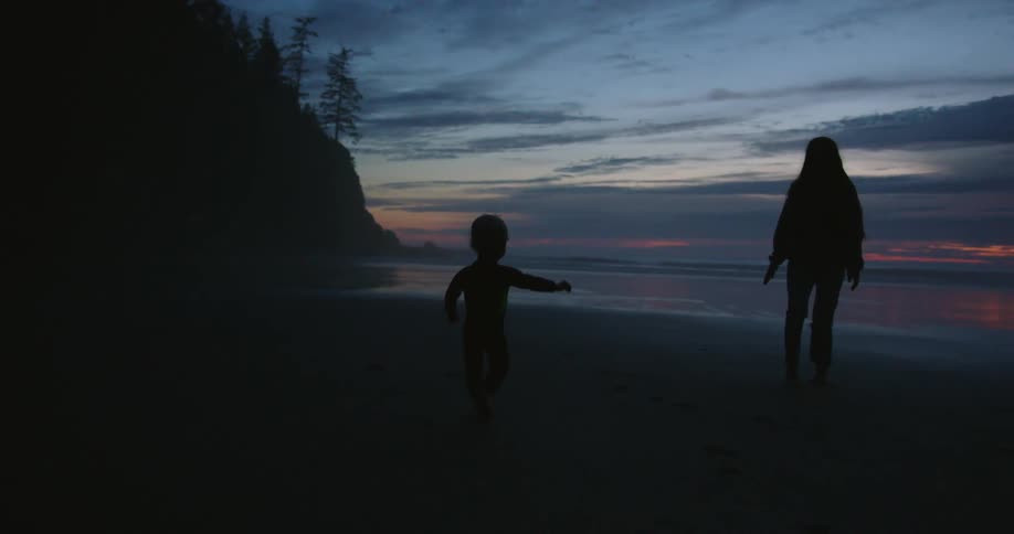 Mother and son walking on beach next to ocean are silhouetted against the red sky at dusk
