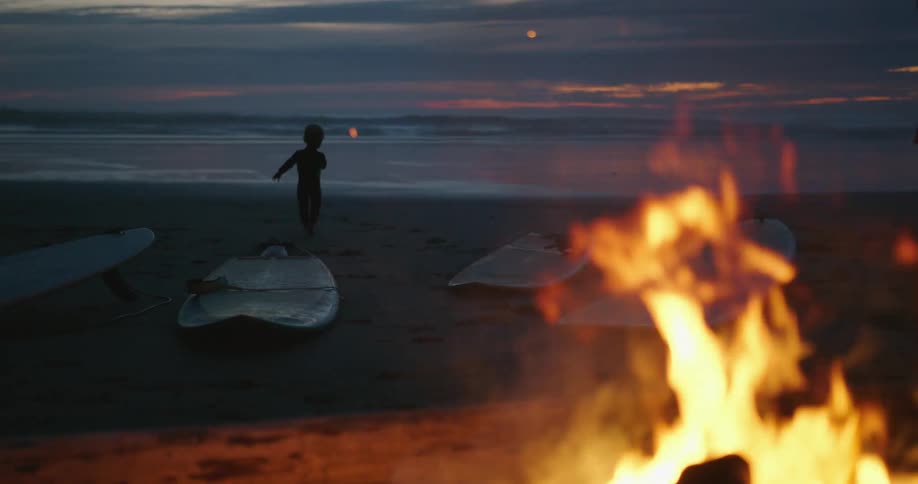 Surfboards are lying on a beach as a bonfire burns nearby and a small child walks near the water
