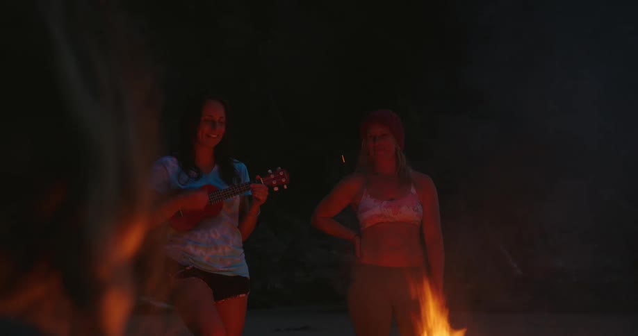 A woman plays the ukulele while standing around a campfire with a group of women at night