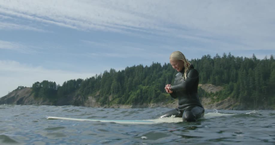 A woman checks the time on her wristwatch while sitting atop a surfboard on the ocean during the day