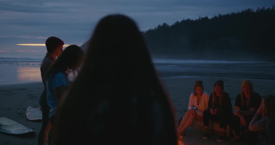 A group of women sit around a campfire by the water