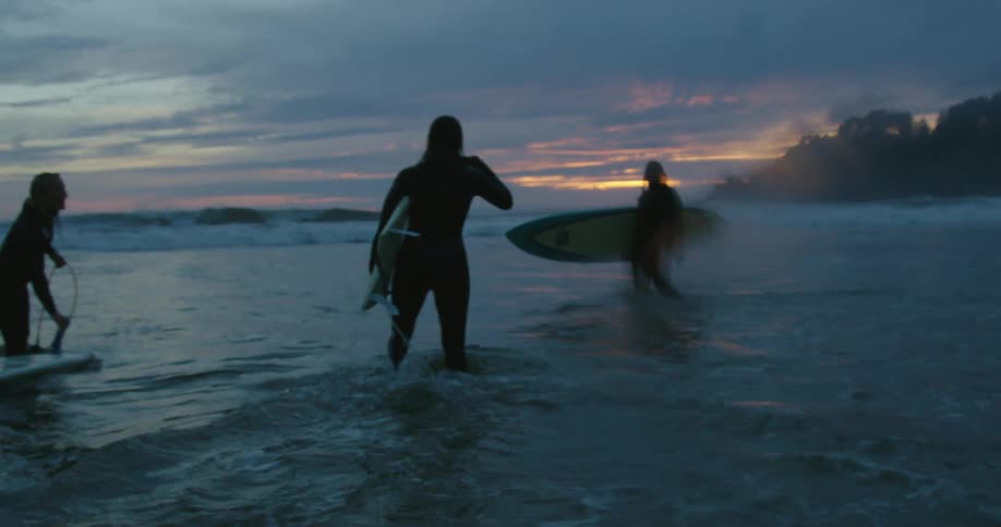 A group of people are wearing wetsuits and carrying surfboards into the water as the sun is setting in a cloudy sky