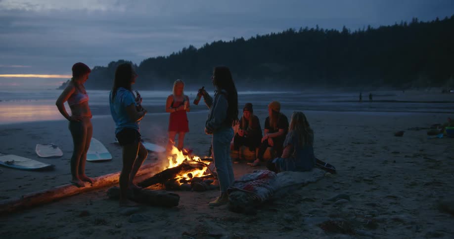 A group of surfers sitting around bonfire talks to each other and drinking beer on the beach at dusk