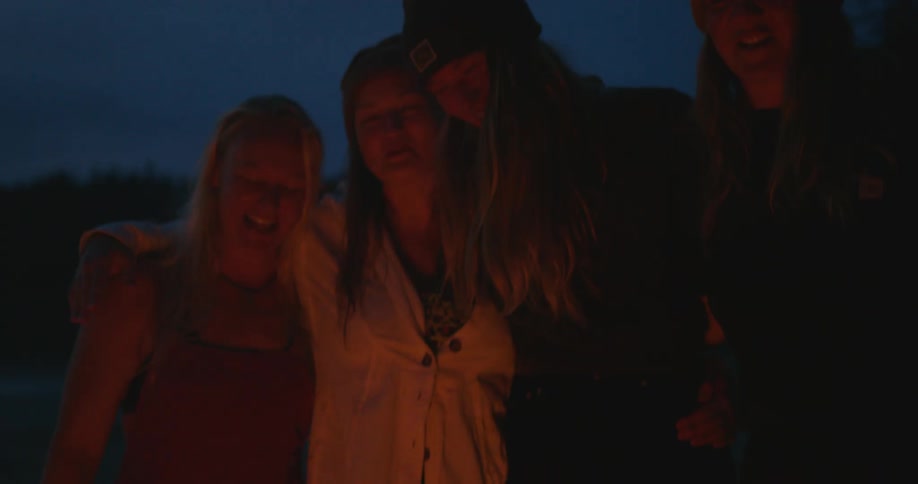Female friends stand around campfire with their arms around each other as they laugh and talk
