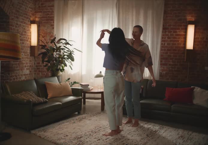 Short hair woman with shirt and jeans holds and dances with woman with long straight hair purple shirt and jeans in living room at home