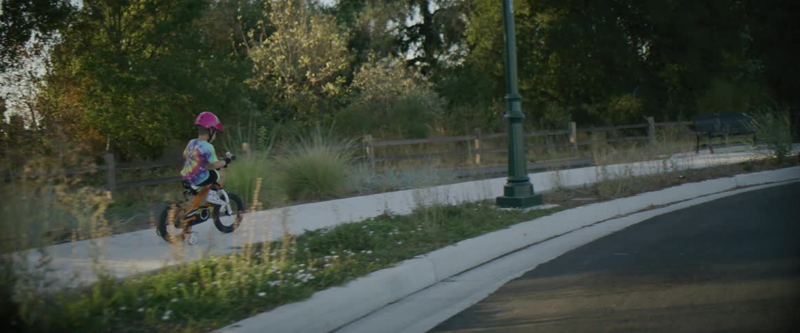 Boy wearing a pink helmet rides a bicycle with training wheels on the sidewalk at morning