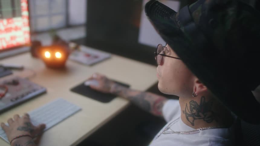 Young man wearing glasses and a hat with tattoos on his neck working on a computer
