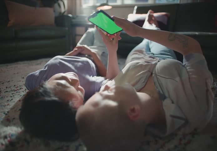 Two people lie on their back on the living room floor sharing a mobile phone screen