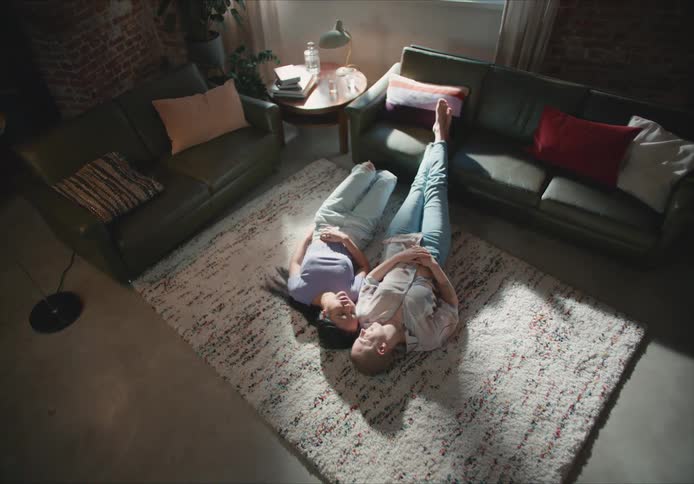 Short hair woman with shirt and jeans lies on the rug with woman with long straight hair purple shirt and jeans talking to each other in living room at home