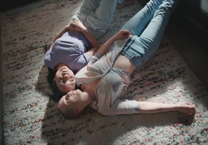 Short hair woman with shirt and jeans lies on the rug with woman with long straight hair purple shirt and jeans talking to each other in living room at home