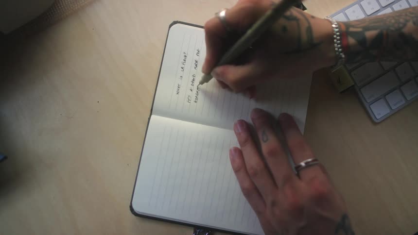 Man with tattoo black hat and glasses writes down in notebook and uses desktop