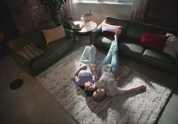Two women lie on the floor in a living room and hold hands