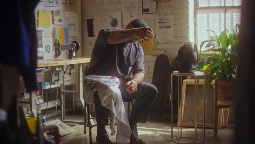 Man sits on stool in sewing room embroidering fabric held in large embroidery hoop