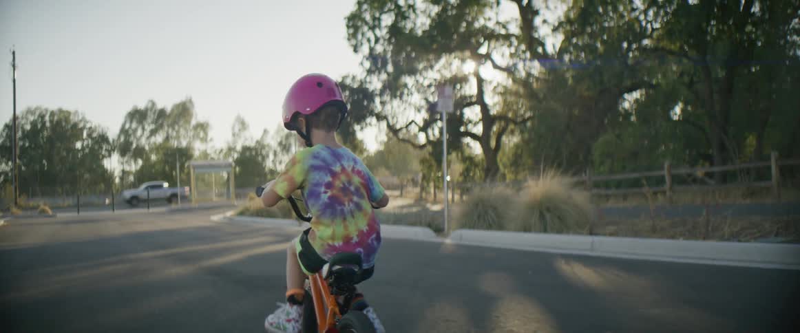 Boy with pink helmet and multi color t shirt shorts and white sneakers cycles on the street in the late afternoon
