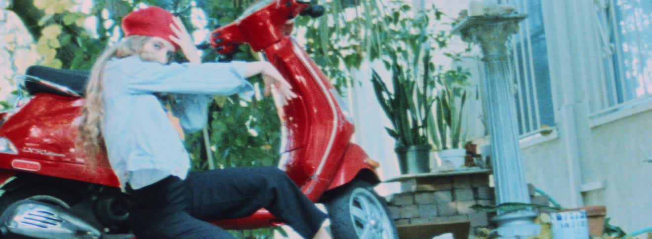 A teenage girl wearing a red had and a light blue jacket is posing next to a red moped
