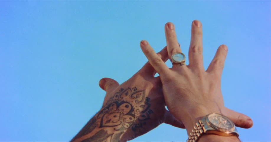 Male hand with tattoos and female hand with wristwatch and ring extending fingers to the blue sky