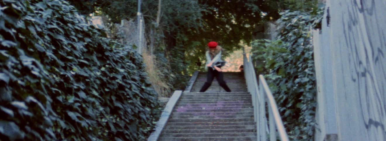 Blonde woman wearing red beret dances on landing of outdoor staircase bordered by walls covered in ivy