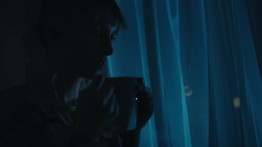 Young woman holding a hot beverage near a window inside a dark room at home