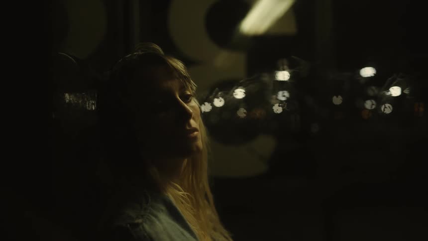 A blonde haired woman is sitting alone in a dark place with lights in the distance and she is looking very depressed