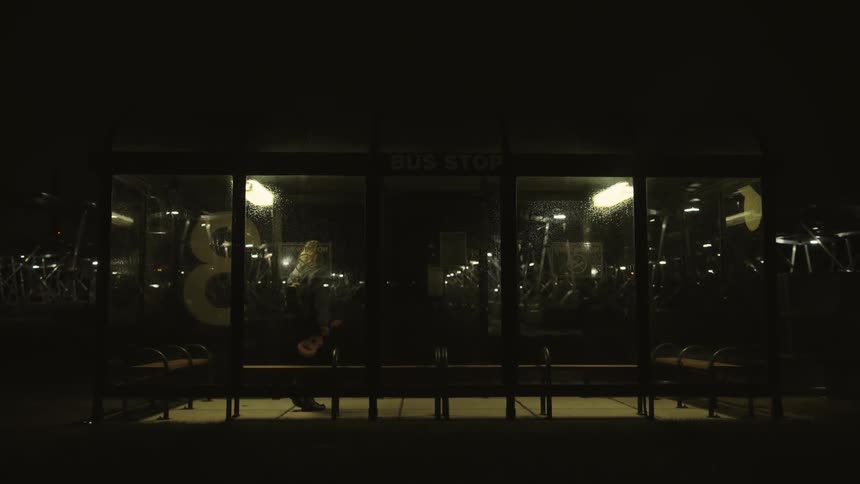 Blonde woman carrying a small musical instrument waiting at a bus stop at night