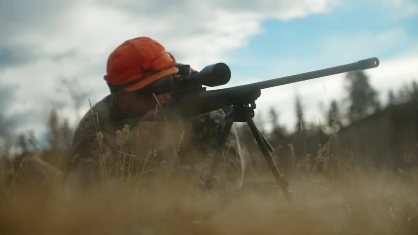 A man crouches in the grass and aims his gun and fires