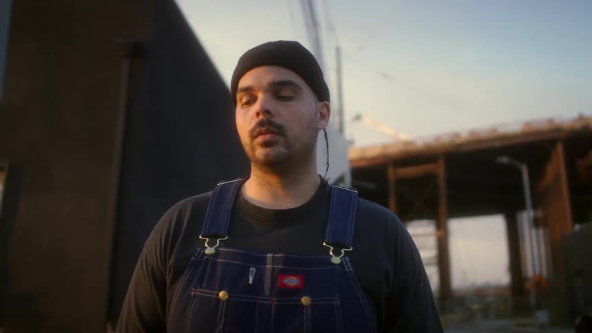Man wearing overalls and a black beanie looks at the camera standing near a construction site
