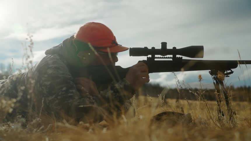 A man wearing an orange hat is lying on the ground looking through the scope of a rifle and taking aim