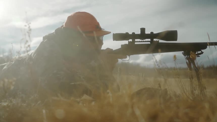 A man wearing an orange hat is lying on the ground looking through the scope of a rifle and shooting
