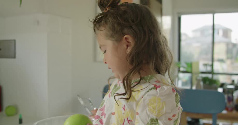 Toddler girl with wavy hair and dress sitting on kitchen counter peels green apple in the kitchen