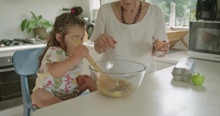 A young girl is in the kitchen with her grandmother stirring a mixture in a bowl with a wooden spoon