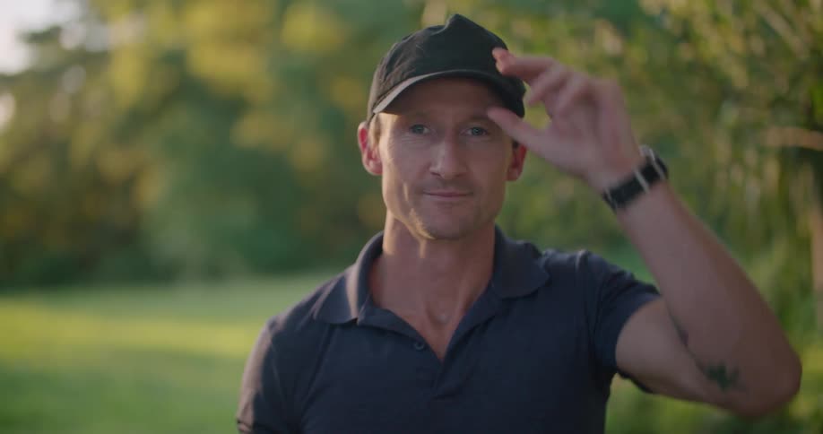 A man standing outside in a field takes off his hearing protection glasses and hat and smiles as he looks straight ahead
