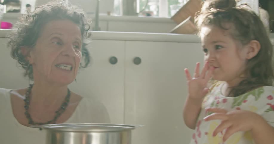 A woman holds a cake pan as she sits on the floor of her kitchen laughing with her grandaughter