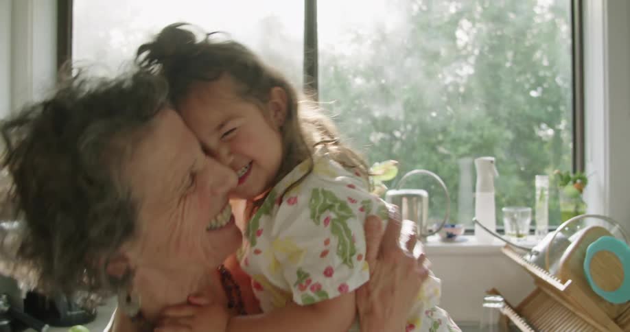 Grandmother stands in kitchen holding granddaughter and they smile as they rub noses together