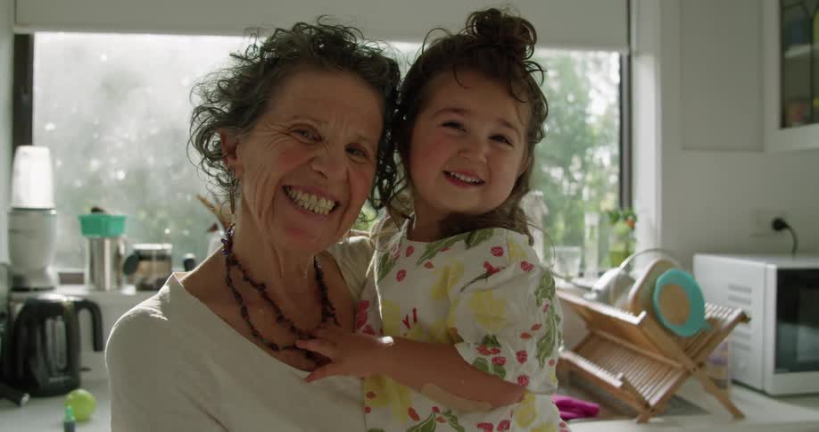 Grandmother holds granddaughter as they smile in kitchen in front of sink and window