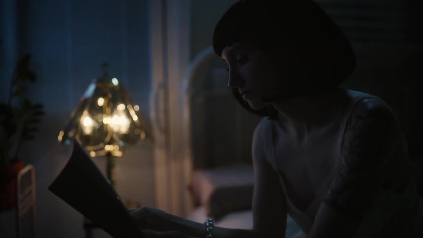 Woman sits in dark bedroom lit by lamp reading