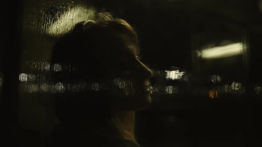 A woman listens to headphones while standing next to a window on a rainy day