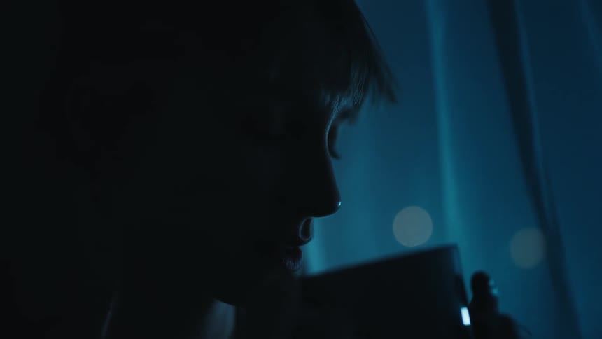 A blonde haired woman is sitting alone in the dark drinking from a cup