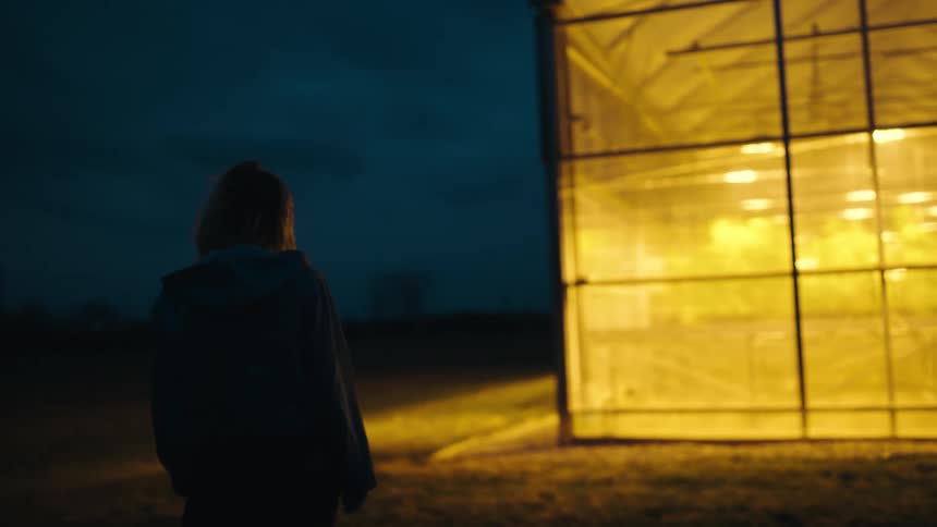 A woman walks past a lit greenhouse at sunset