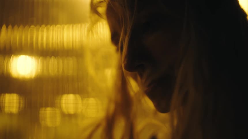 Blonde woman back lit by amber glow looks down as windy blows long hair over her face