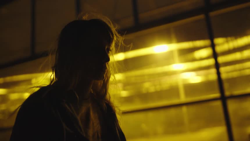 A blonde haired woman is walking outside at night past buildings with bright lights on inside