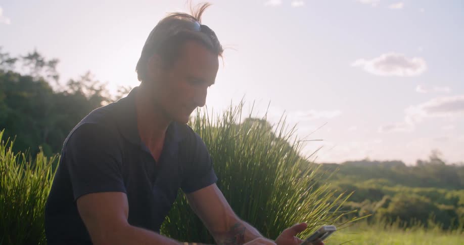 Man sitting on the ground near a grass field looking at his phone in the morning