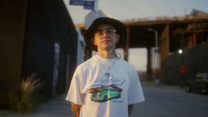 Man wearing hat and eyeglasses stands near street with overpass in background