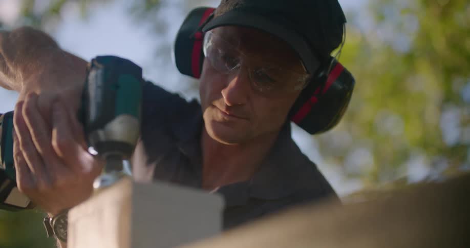 A man wearing ear muffs is sanding the top of a fence post
