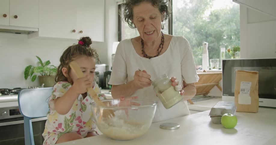 A toddler girl helps her grandmother mix ingredients in a home kitchen