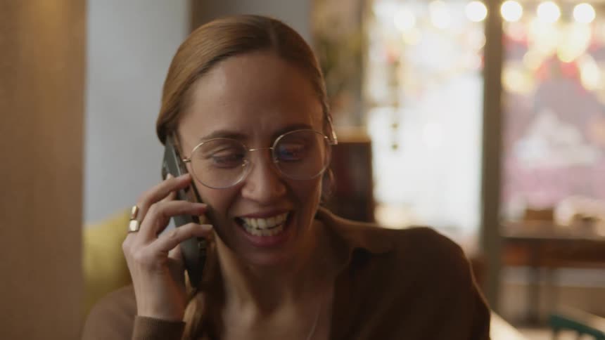 Woman with brown hair sweater glasses rings and hoop earrings talks on smartphone smiling in cafe