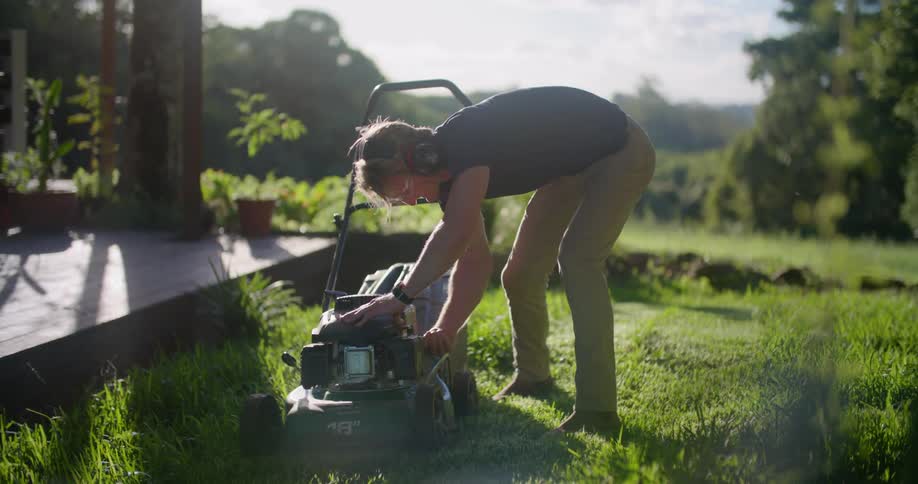 Man with black t shirt and brown pants starts lawn mower to mow grass next to country house