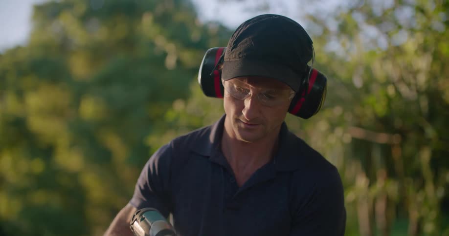 A man wears protective earmuffs and goggles while sanding a fence in his yard