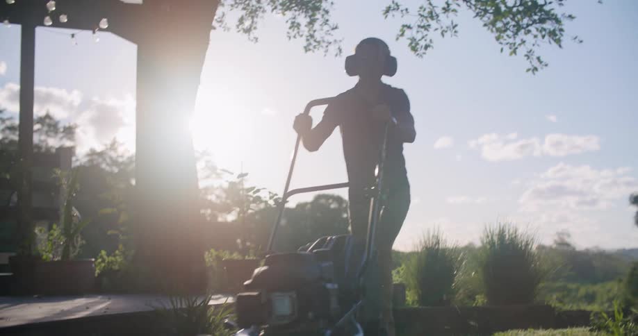 Man wearing earmuffs uses a lawnmower on the backyard at morning
