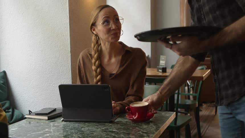 Woman working on a laptop at a restaurant is served a red cup of coffee