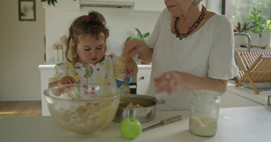 Grandmother with necklace and white shirt makes food next to granddaughter in the kitchen at home